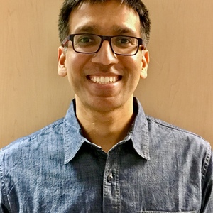 Headshot of researcher wearing glasses and blue button up with a plain beige background